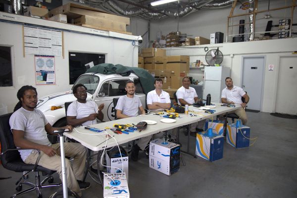 A group of six people sitting at a table in a workshop, surrounded by tools and a vintage car.