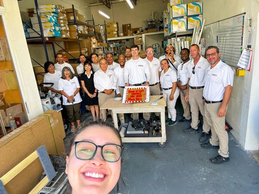 A group of team members posing for a celebratory photo with a cake in a warehouse setting.