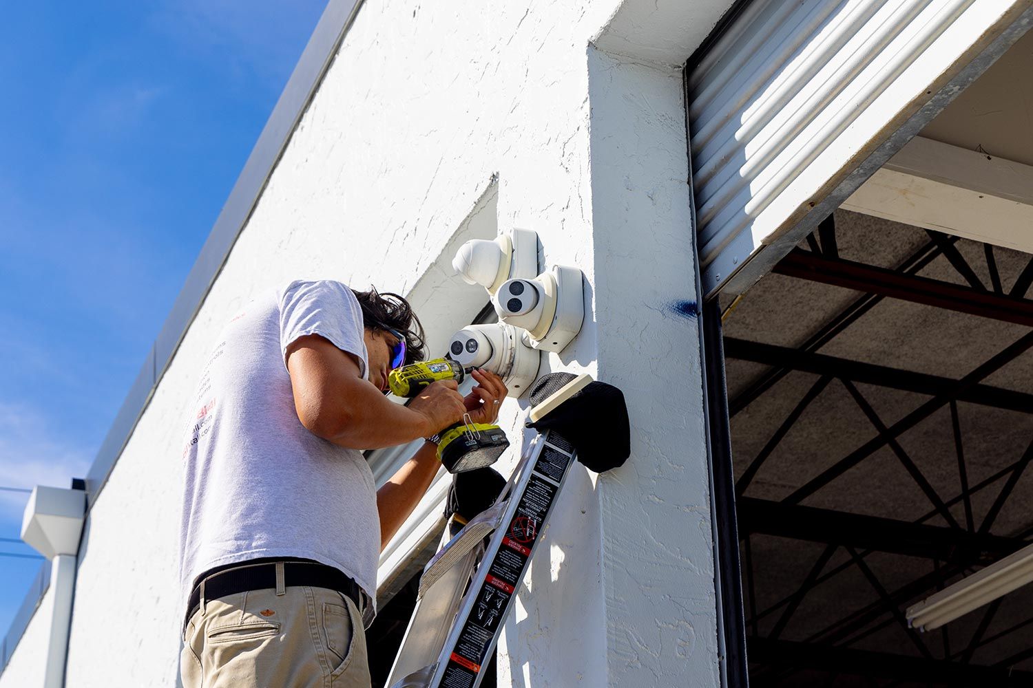 A technician installing outdoor surveillance cameras on a white wall near a warehouse door.