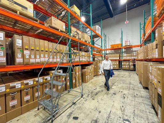 A man walking through a large warehouse with rows of stacked boxes and a rolling ladder nearby.