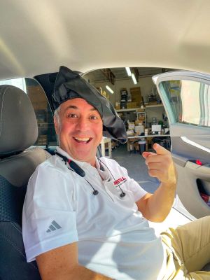 A man sitting in a parked car, smiling and wearing a black cap in a humorous manner.