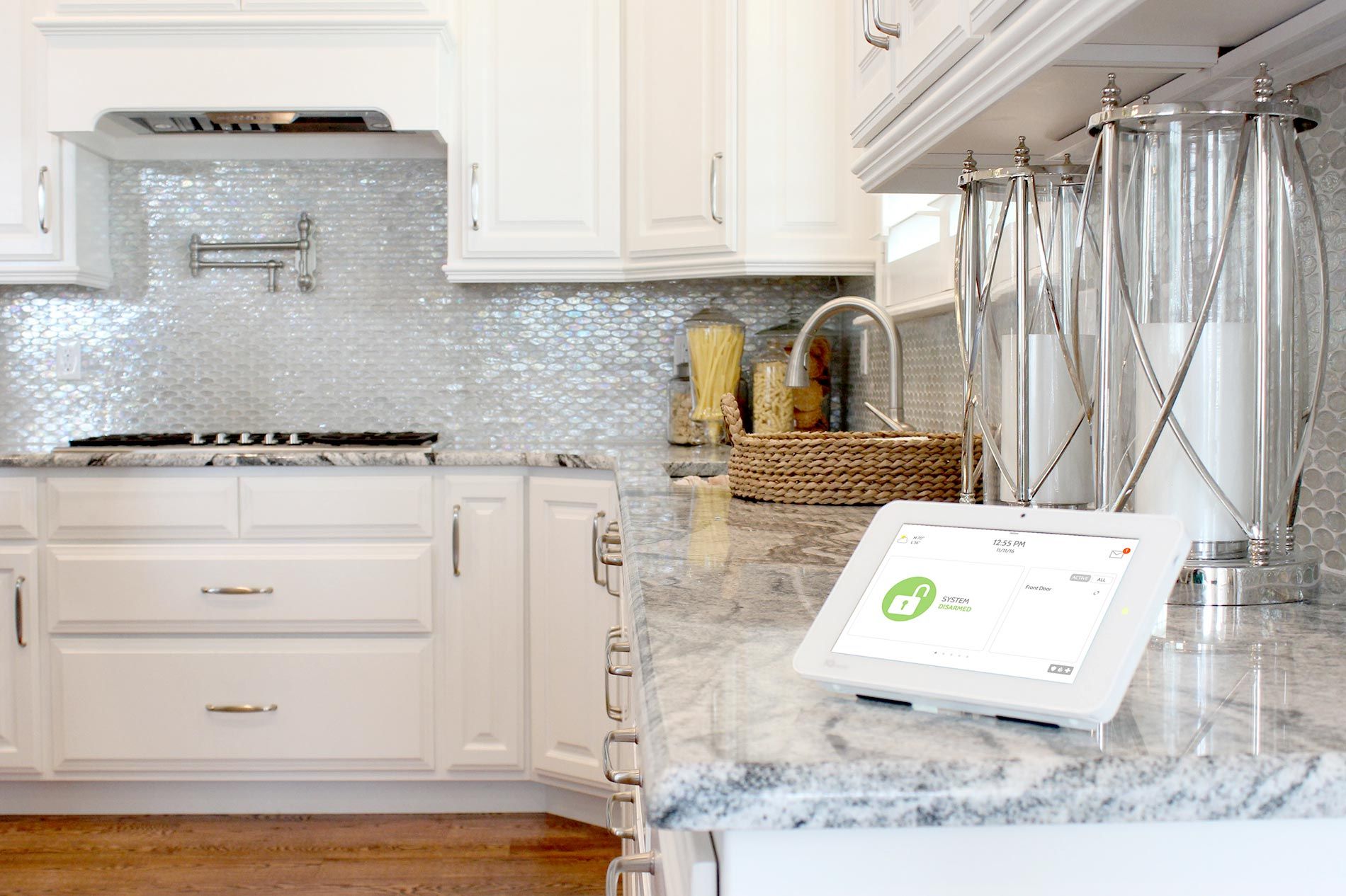 A modern kitchen with a security system tablet on the countertop, featuring sleek white cabinets and a decorative backsplash.