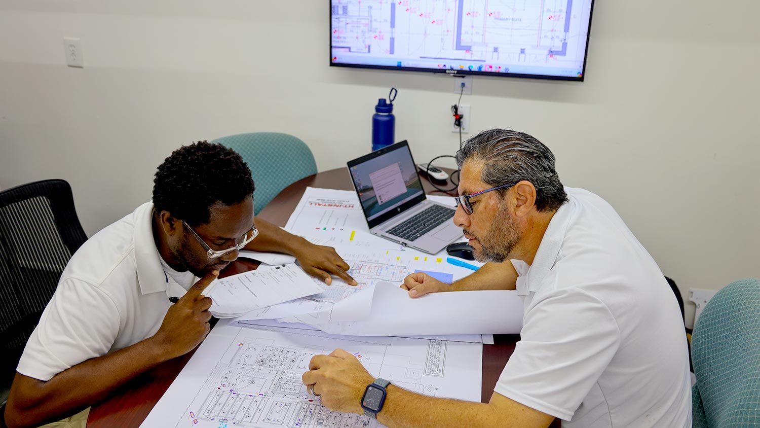 Two colleagues examining a set of blueprints and documents at a meeting table in a planning session.