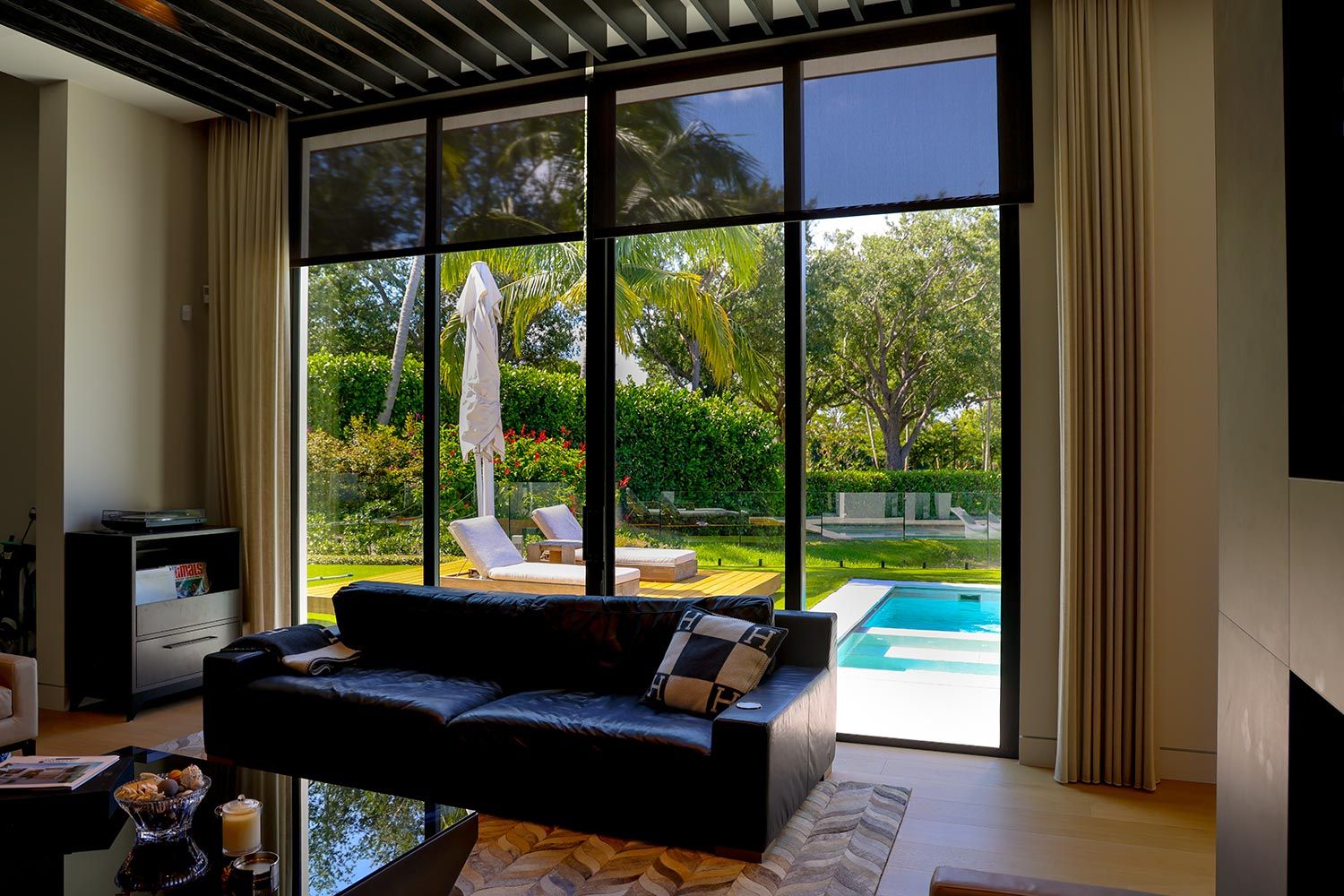 A modern living room with large glass doors leading to a poolside patio, featuring outdoor lounge chairs and lush greenery.